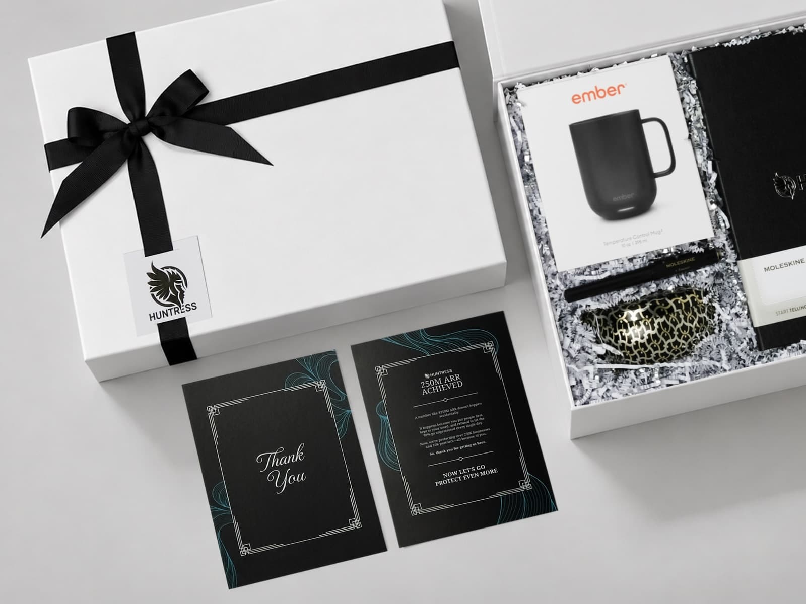 A gift box tied with a black ribbon and a logo tag, thank-you cards and an open box with a mug