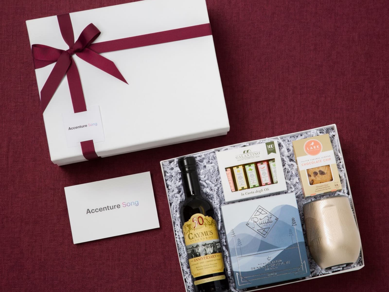 A gift box for Accenture Song with a burgundy ribbon and logo tag, its logo card, and the open box of gifts